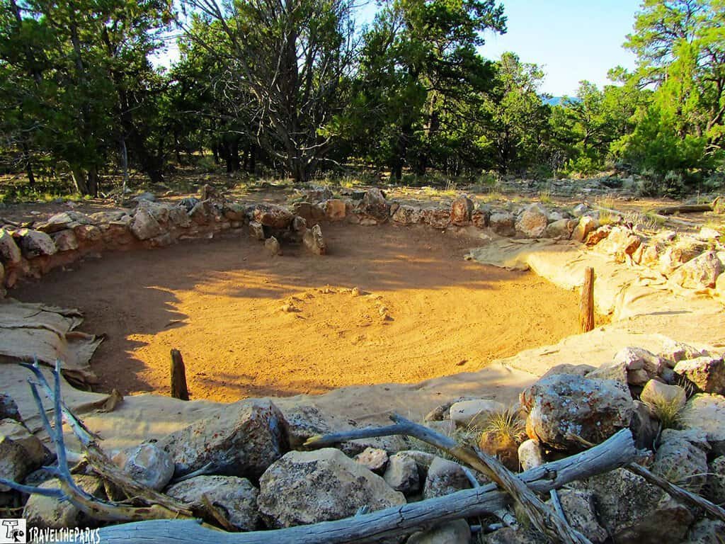 Tusayan Ruin Large Kiva: A circular, ancient stone structure sits in a forest clearing. The sunlit earthen floor is surrounded by rugged rocks, with trees in the background. Peaceful atmosphere.