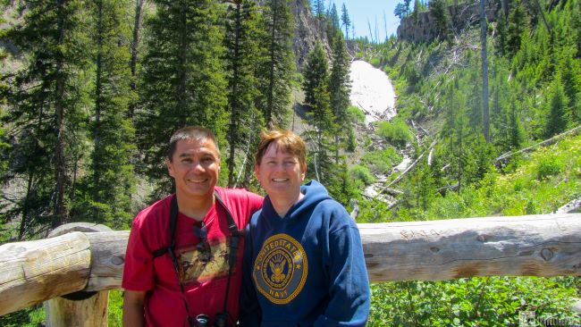 A couple standing in front of a lush forest and Wraith waterfall in Yellowstone NP.