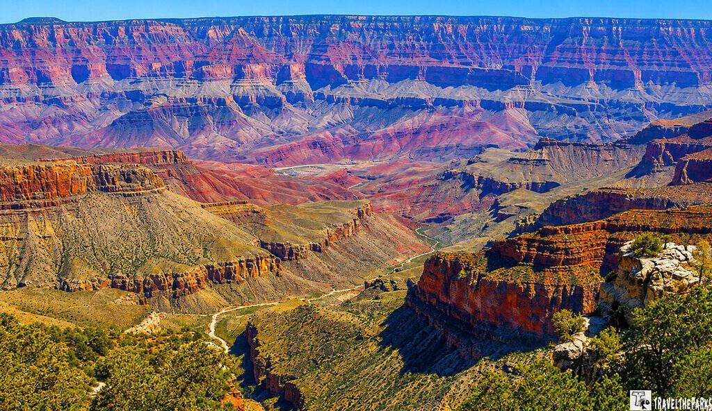 Explore Imperial Point: Vast Grand Canyon landscape with layered red and purple rock formations, deep valleys, and rich greenery, under a clear blue sky. Majestic and awe-inspiring.