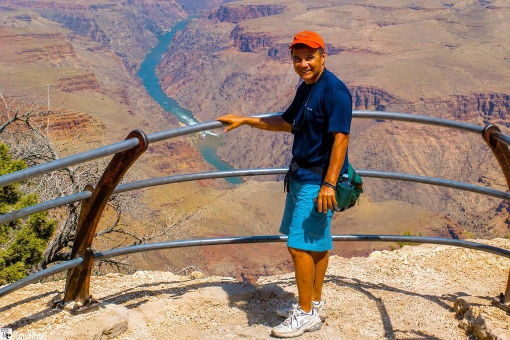A person stands smiling by a railing at the edge of a canyon with a turquoise river below. They wear a red cap, dark shirt, and shorts. It conveys excitement and adventure.