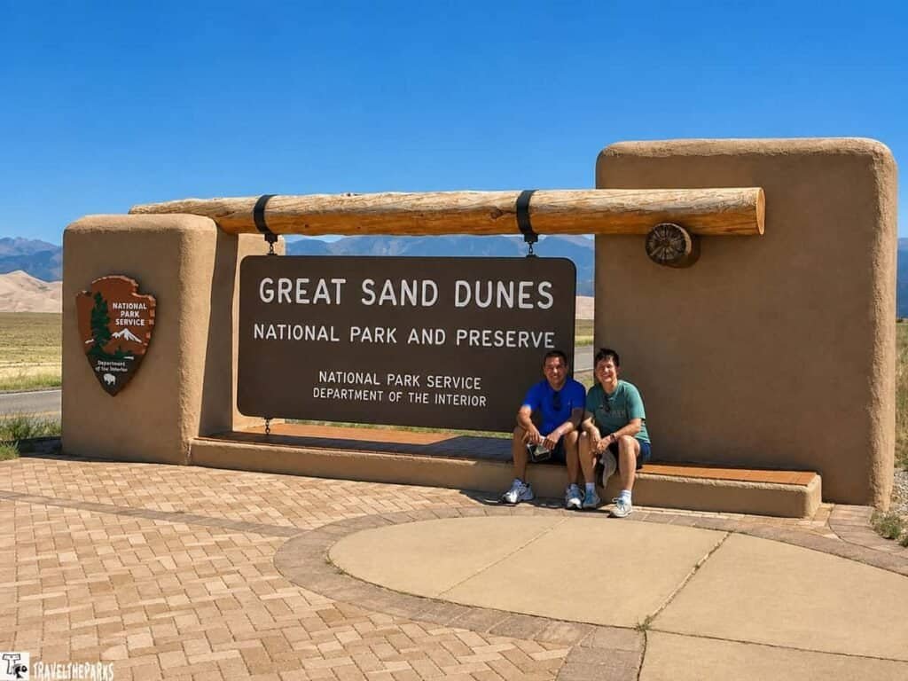 Sign for Great Sand Dunes National Park and Preserve with seating area, set against a plain and mountain backdrop under a clear sky