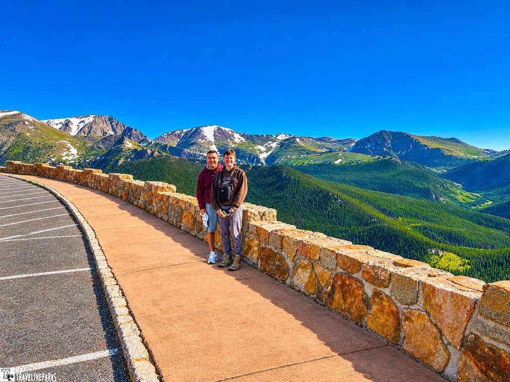 Rainbow Curve, Trail Ridge Road, RMNP: A couple stands on a mountain viewpoint with scenic snowy peaks and vast green valleys under a clear blue sky, conveying a sense of adventure and tranquility.