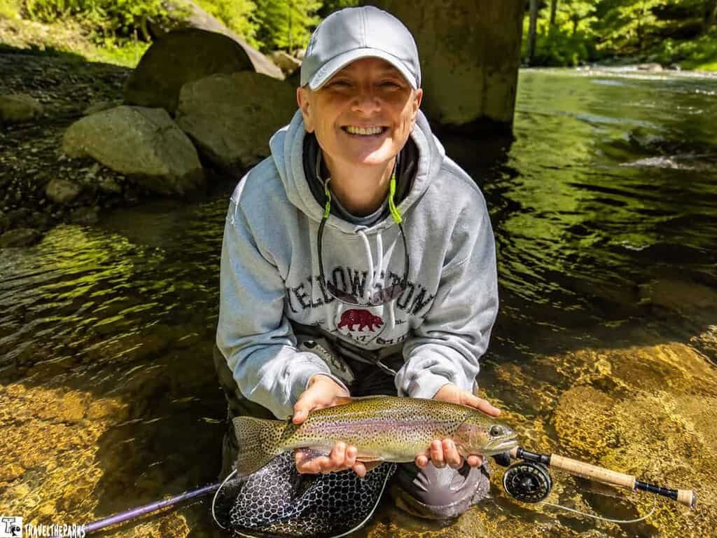 Person kneeling in a stream holding a fish and a fishing rod, wearing a Yellowstone hoodie and a cap.