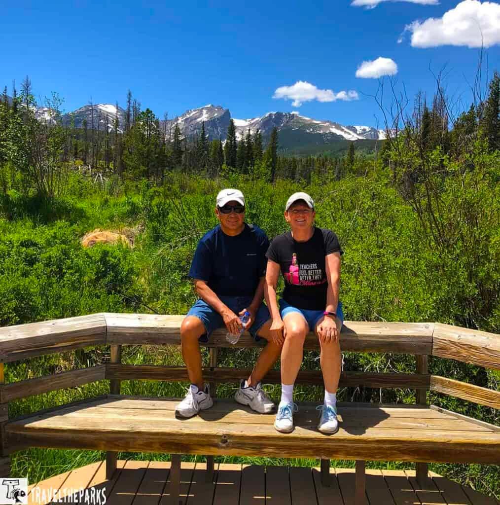 Rocky Mountain NP: Two people sitting on a wooden bench with mountains in the background.