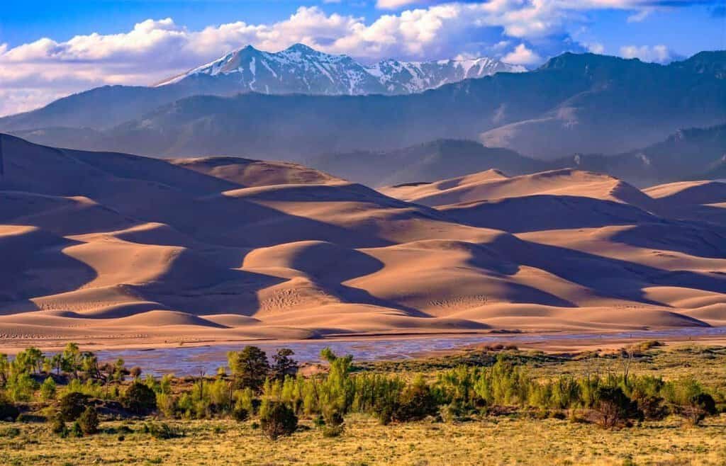 A landscape view showing grasslands, Medano Creek, undulating sand dunes, and snow-capped mountains under a blue sky with clouds.