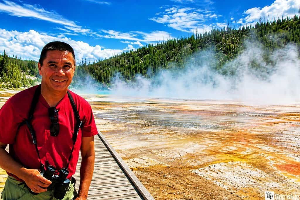 Grand Prismatic Boardwalk: Man in a red shirt smiles holding binoculars at a steamy, colorful hot spring. Blue sky and forested hills in the background suggest a natural setting.