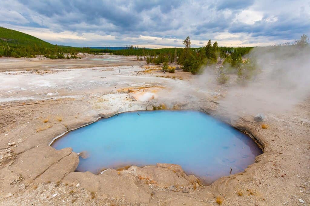 A blue geothermal hot spring surrounded by gray mineral deposits and steam, with a forested hillside and cloudy sky in the background.
