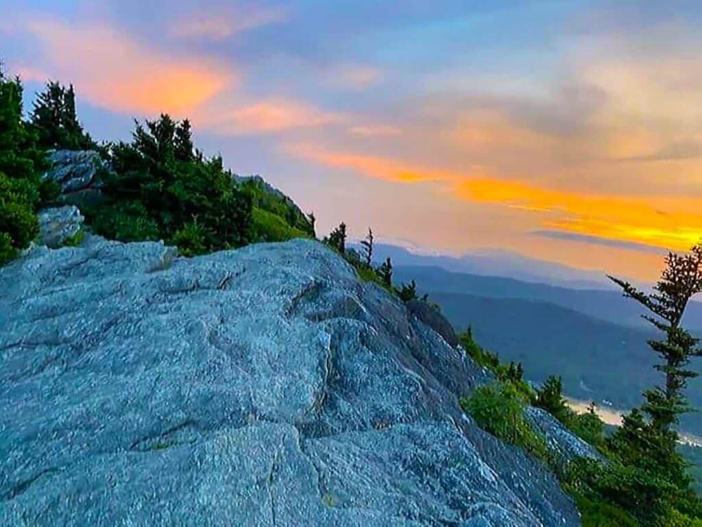 Mountain landscape at sunset with rocks, trees, and colorful sky.
