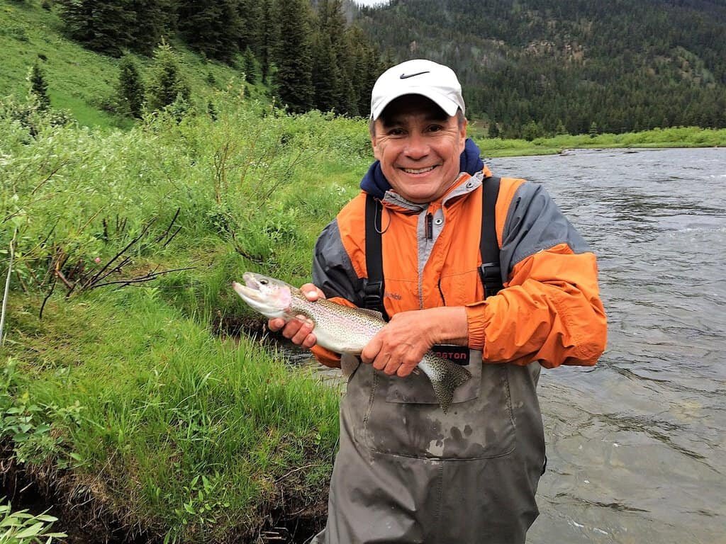 Madison River A smiling person in an orange jacket and cap holds a fish by a river with lush green foliage and trees in the background, conveying joy and success.