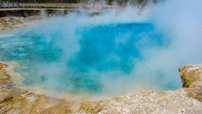 Excelsior Geyser: A vibrant blue hot spring with steam rising, surrounded by rocky terrain. A wooden railing is visible in the background, evoking a serene, natural wonder.