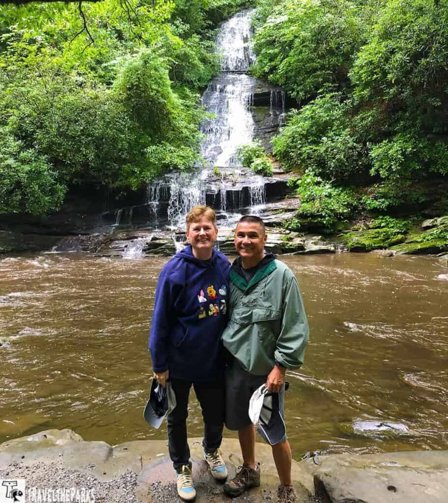 a tranquil outdoor setting featuring a waterfall cascading down a rocky, forested landscape. The waterfall is tiered, meandering down smooth stone layers surrounded by lush greenery. In the foreground, two individuals stand on a rocky edge by a calm, brown-hued river. They are dressed in casual, outdoor clothing, each holding a hat. The area is enveloped with dense vegetation, highlighting the natural beauty of the scene. Sunlight filters through the leaves, casting dappled light on the waterfall and water surface.