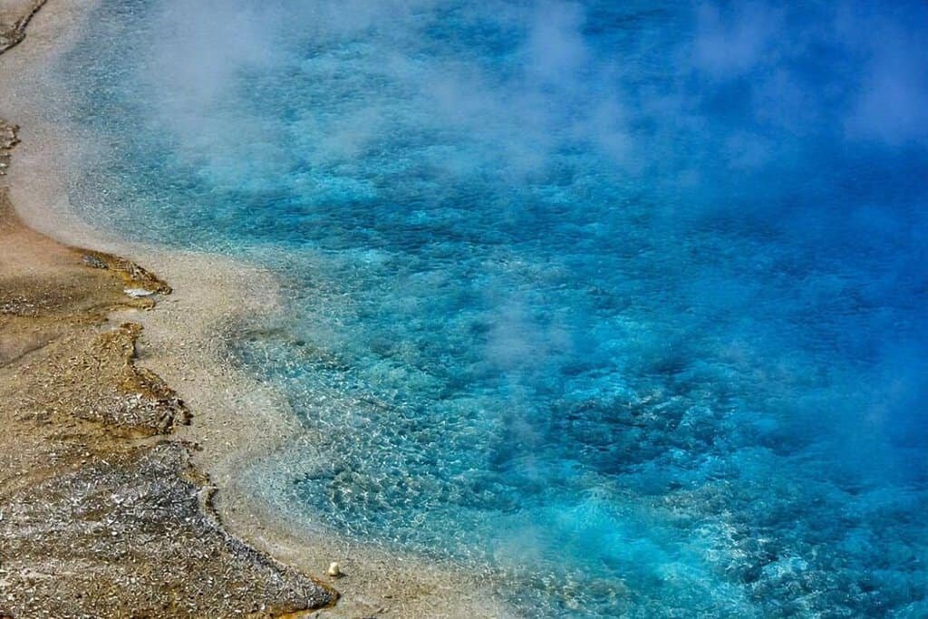 Excelsior Geyser: A vibrant blue hot spring with steam rising, surrounded by rocky terrain. A wooden railing is visible in the background, evoking a serene, natural wonder.