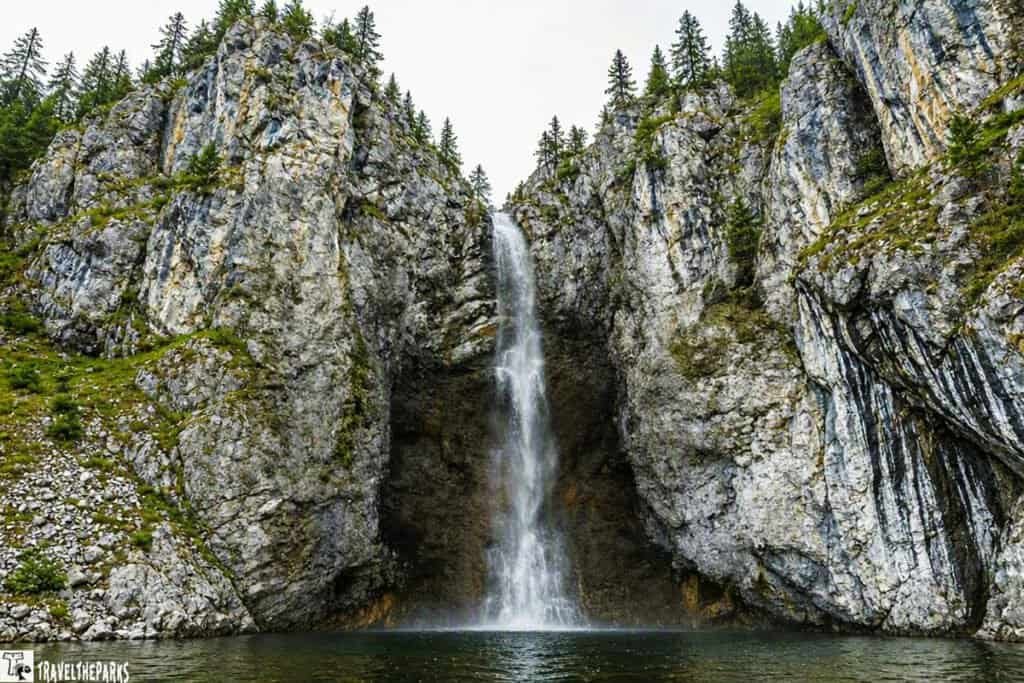 A tall fairy waterfall cascades down a rugged, rocky cliff into a calm pool below, creating a serene and natural scene. The rocks are textured and gray, complementing the clear, flowing water.