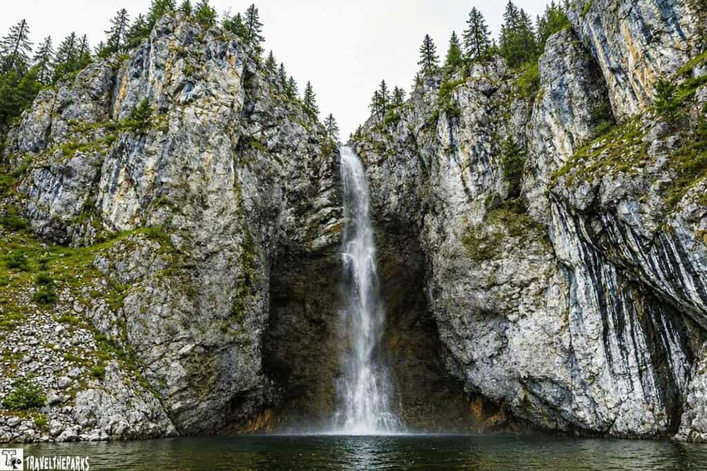 A tall fairy waterfall cascades down a rugged, rocky cliff into a calm pool below, creating a serene and natural scene. The rocks are textured and gray, complementing the clear, flowing water.