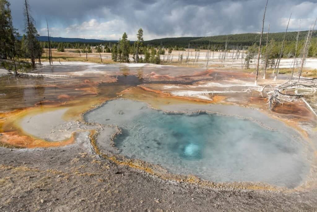 A circular turquoise geothermal spring surrounded by gray and orange mineral deposits, with a backdrop of trees and a cloudy sky in the Lower Geyser Basin.