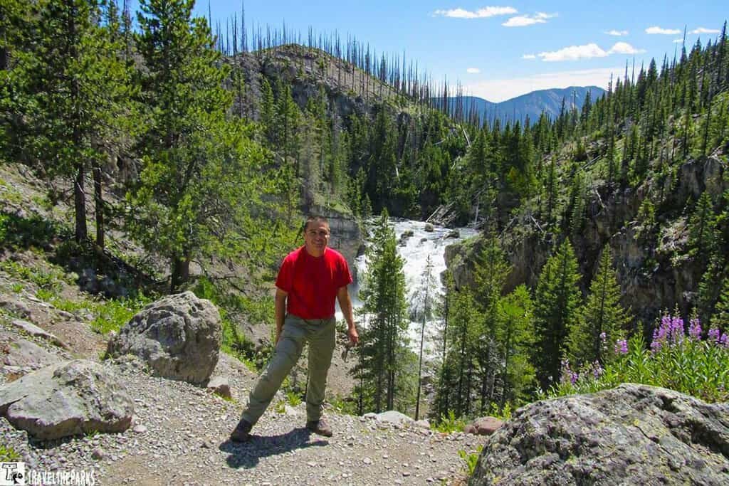 FIREHOLE SWIMMING HOLE-Yellowstone NP: A person in a red shirt smiles on a forested trail next to a waterfall. Pine trees and bright sunlight create a serene, nature-focused atmosphere.