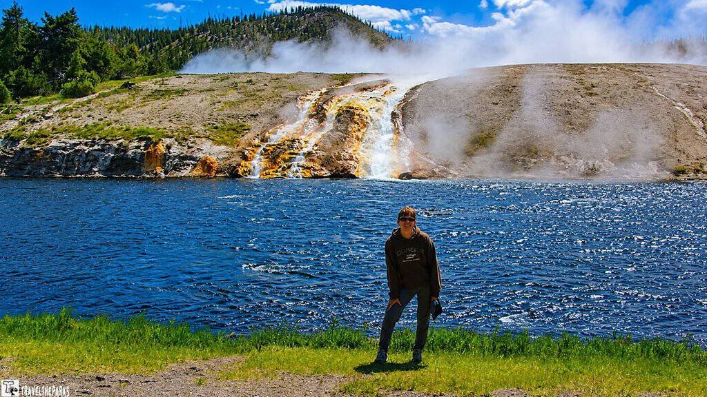A steaming geyser flows into a river under a blue sky, surrounded by lush greenery and pine trees, creating a serene and dynamic natural scene.
