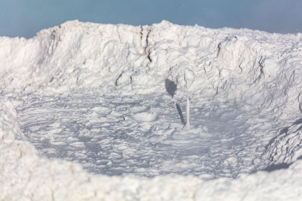 A bubbling mud pot with white, textured mud and a blue sky in the background, featuring splashes of mud from bursting bubbles.