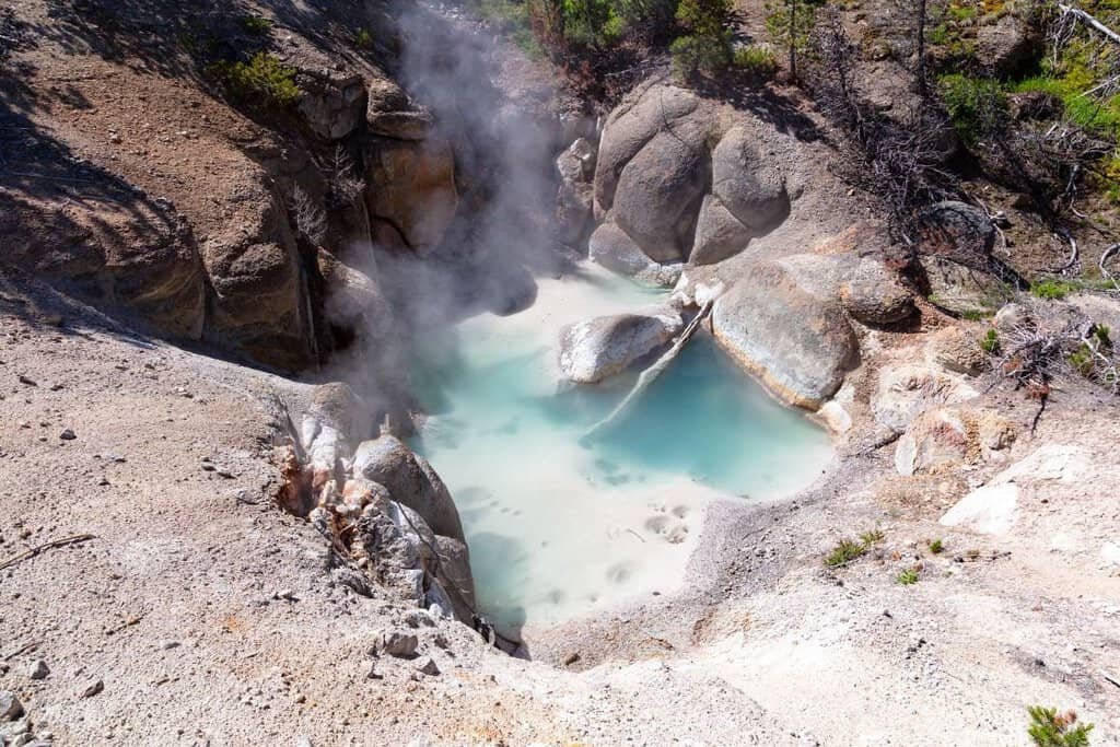 Geothermal pool surrounded by rocky terrain and steam rising in the Norris Geyser Basin, showcasing blue water and textured rocks.