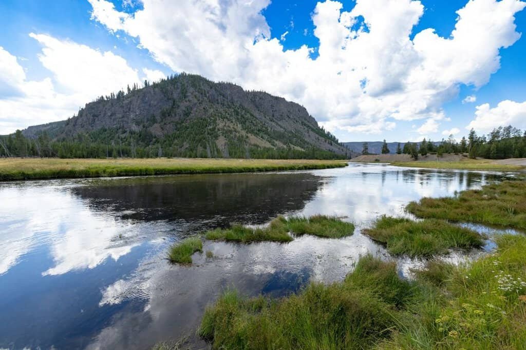 A scenic view of the Madison River in Yellowstone, featuring flowing water, lush green banks, and a rocky hill in the background under a blue sky with clouds.