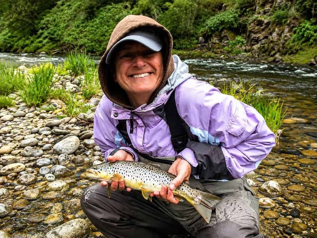 Smiling person in a hooded jacket proudly holds a brown trout by a rocky riverbank. The background features lush greenery and flowing water.