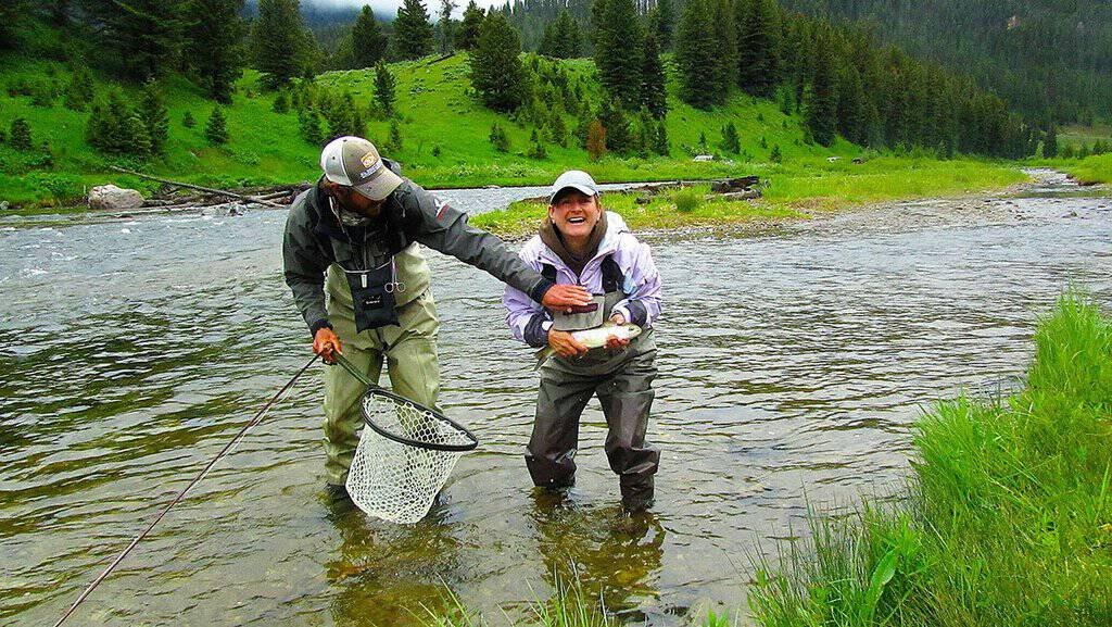 Smiling person in a hooded jacket proudly holds a brown trout by a rocky riverbank. The background features lush greenery and flowing water.