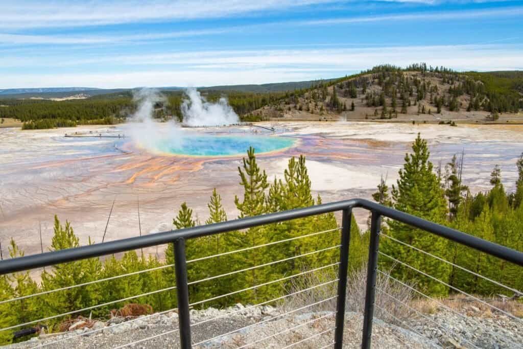 A view from the Grand Prismatic Overlook Trail shows the colorful Grand Prismatic Spring surrounded by steam and a metal railing in the foreground.