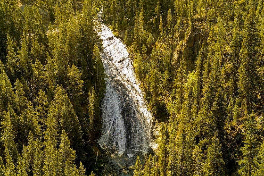 A waterfall cascading down a rocky slope, surrounded by dense green trees, with sunlight reflecting off the water.