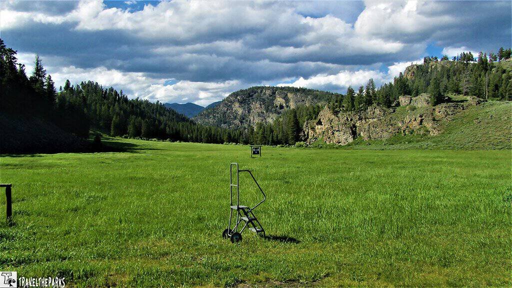 Wagon Train Dinner-Yellowstone National Park: A lush, green grassy field under a partly cloudy sky stretches toward distant forested hills and mountains. A step ladder stands in the foreground, creating a whimsical contrast with the serene landscape.