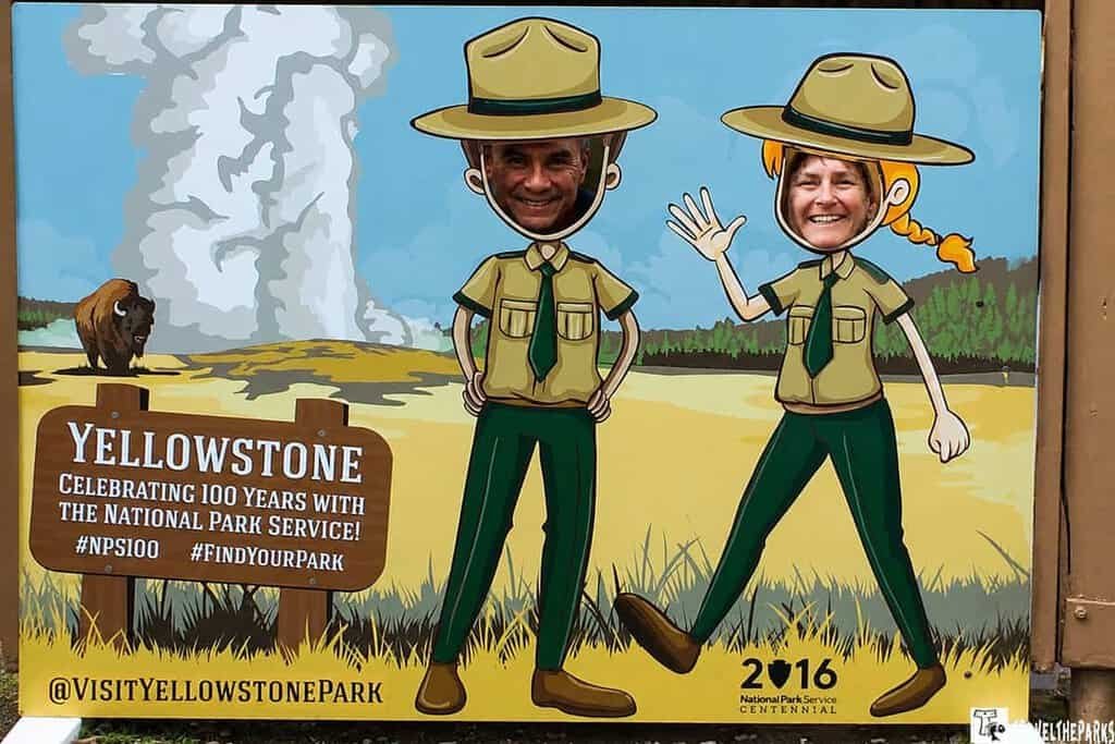 Two people smile through a cutout display of cartoon park rangers at Yellowstone, celebrating the National Park Service's centennial.