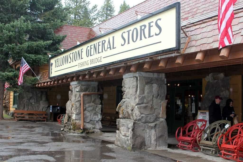 Exterior of Yellowstone General Stores at Fishing Bridge, featuring a prominent sign, stone pillars, and people standing at the entrance under a cloudy sky.