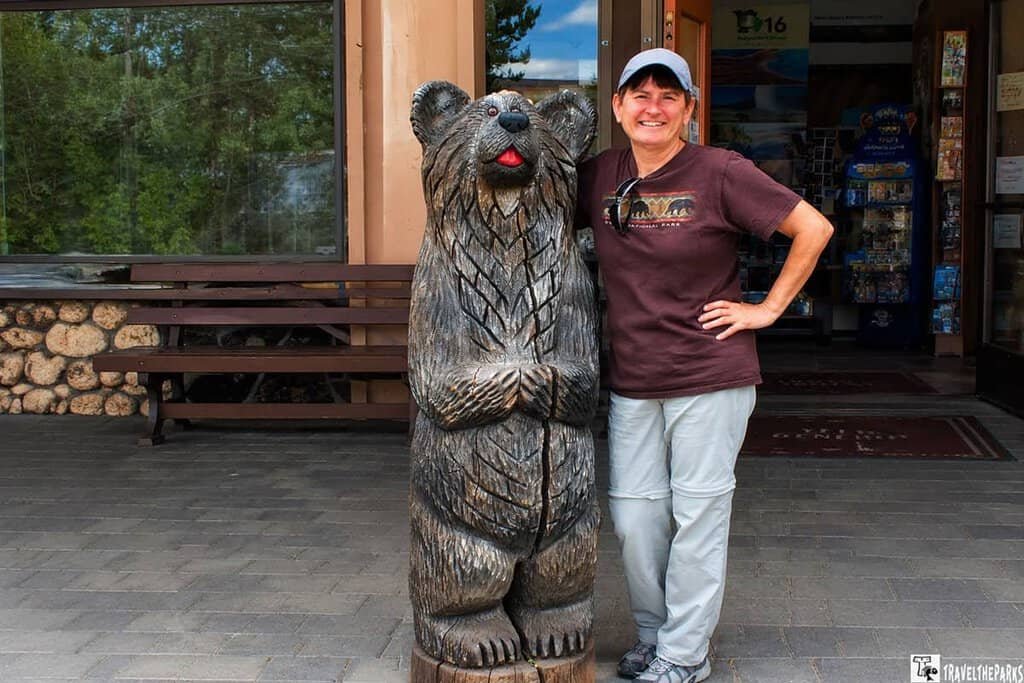 A woman smiles next to a wooden bear sculpture at Canyon Village in Yellowstone National Park, with a rustic building and green trees in the background.

