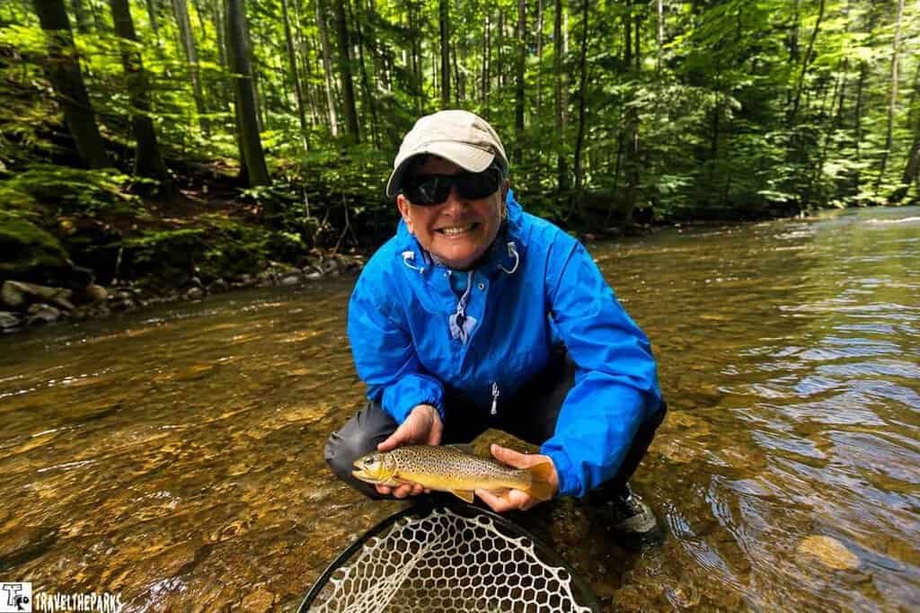 A smiling person kneeling at the riverbank holds a brown trout beside a fishing net, with lush green forest in the background.

