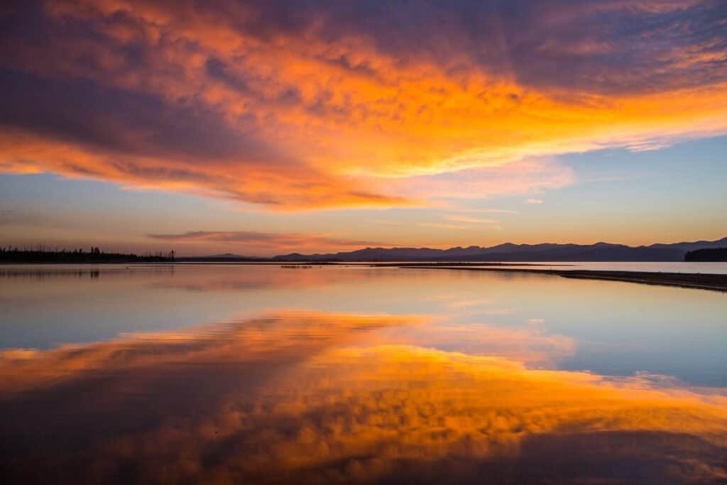 A vibrant sunrise over Yellowstone Lake, with reflections of orange and purple clouds on the calm water and distant mountains in the background.