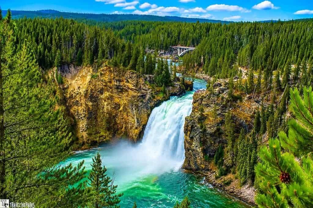 A waterfall cascades down a rocky cliff into a turquoise river, surrounded by evergreen trees and a bridge in the background under a blue sky with clouds.

