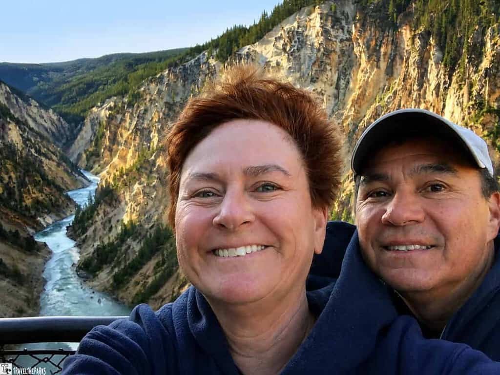 Two people take a selfie with the Grand Canyon of Yellowstone in the background, showcasing steep cliffs and a winding river.
