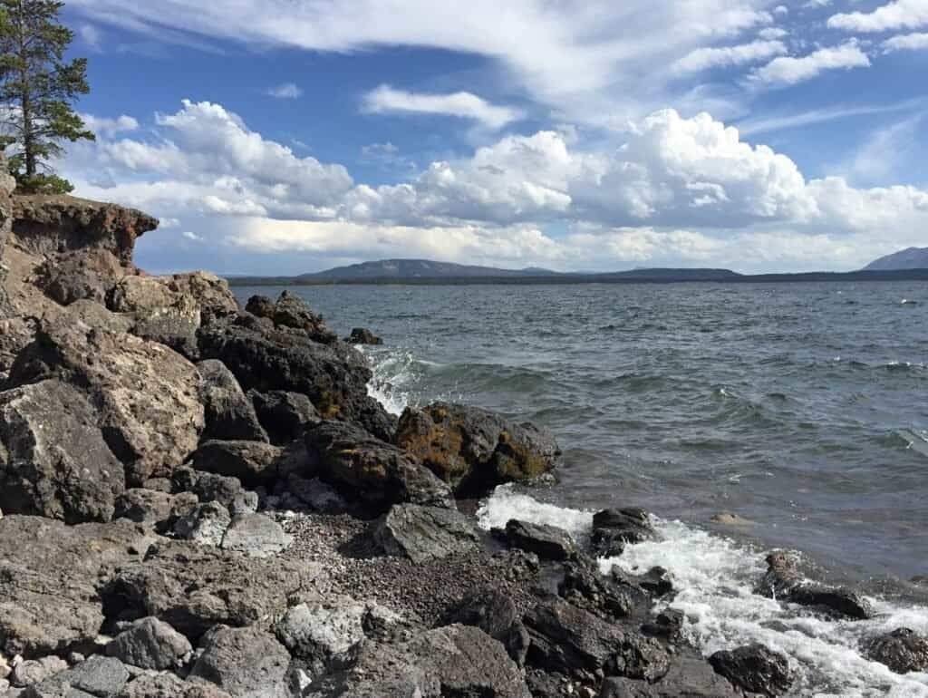 A rocky shore at West Thumb Lake features dark volcanic rocks and gentle waves. A tree is visible on the left, with mountains and a partly cloudy sky in the background.