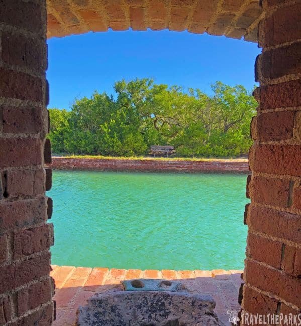 Dry Tortugas-Fort Jefferson-Looking out: A view of a calm green waterway framed by brick walls, with lush greenery in the background under a clear blue sky.