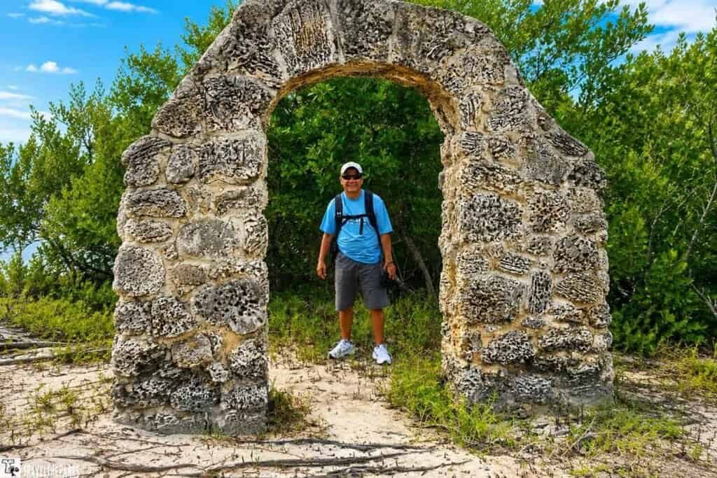 A weathered stone archway surrounded by green vegetation on Boca Chita Island, with a person in light blue attire standing in the center.


