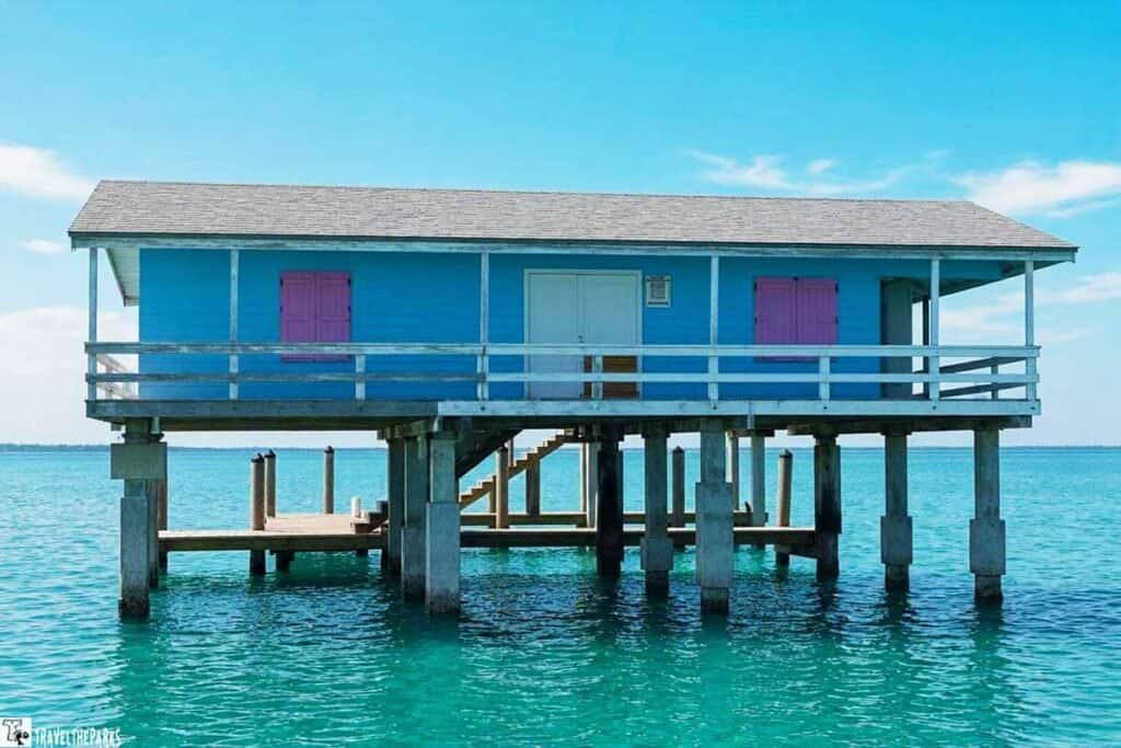 A brightly colored blue stilt house with pink shutters above turquoise water, featuring a wooden deck and stairs.