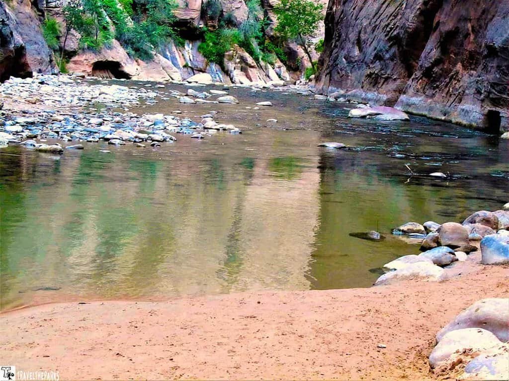 A serene view of The Narrows at Zion National Park, featuring sandy banks, scattered stones, and gently flowing water reflecting canyon walls and greenery.

