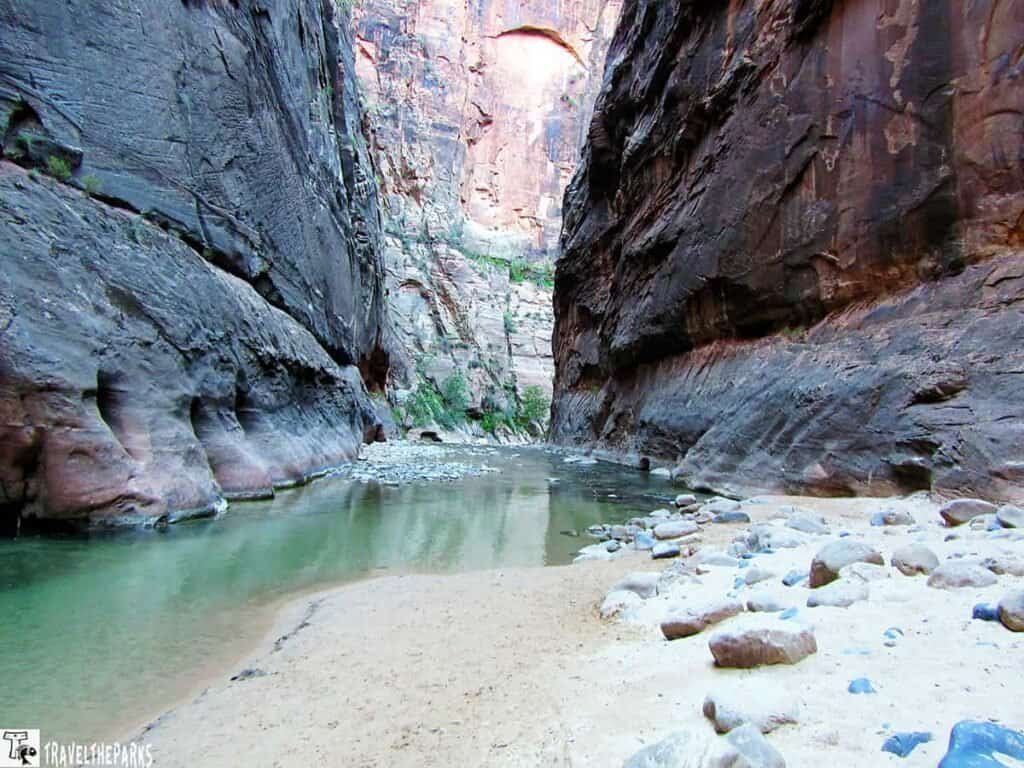 The-Narrows-Temple-of-Sinawava: Narrow canyon with towering rock walls and a shallow river winding through sandy terrain. The scene conveys a sense of solitude and natural beauty.