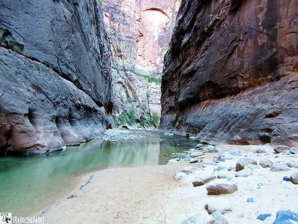 The-Narrows-Temple-of-Sinawava: Narrow canyon with towering rock walls and a shallow river winding through sandy terrain. The scene conveys a sense of solitude and natural beauty.