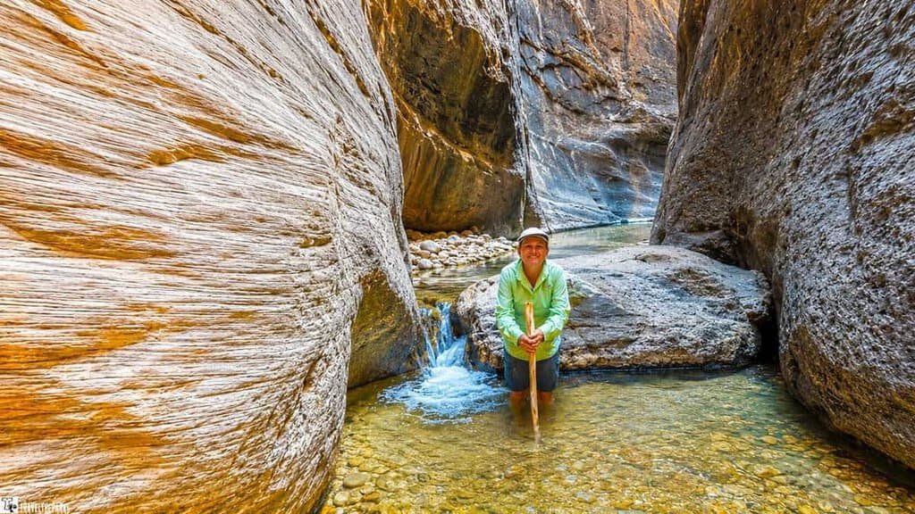 A person in a green shirt and shorts stands in a shallow stream within a narrow canyon, smiling and holding a walking stick, with rocky walls on either side.

