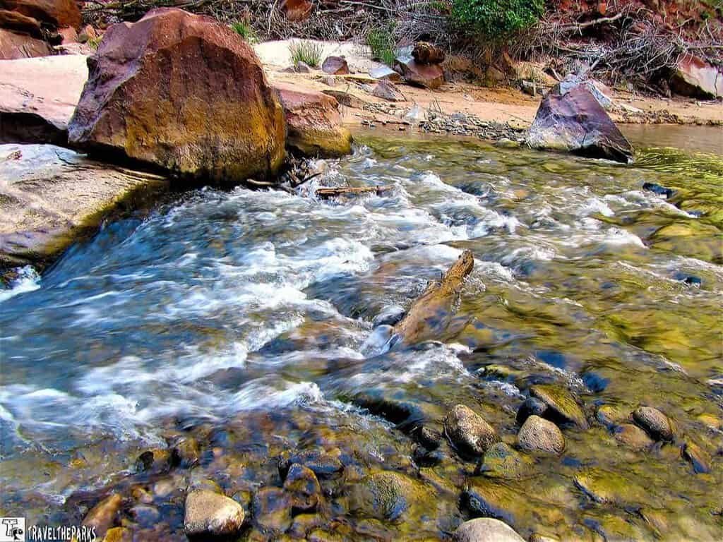 A flowing river with smooth stones and surrounding boulders in shades of brown and red, set in a natural landscape.

