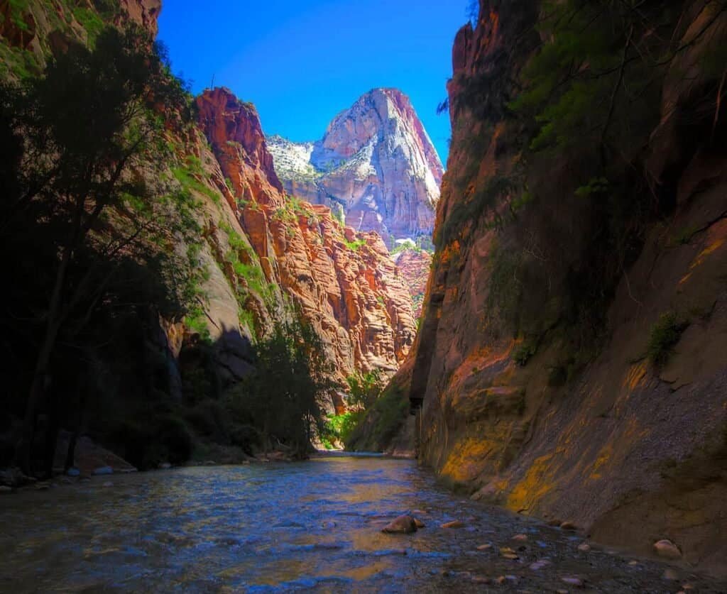 A narrow canyon with colorful rock walls and a river flowing through it, under a clear blue sky. A prominent mountain peak is visible in the background.

