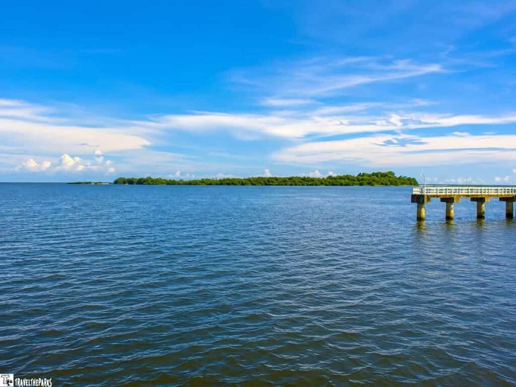 A coastal view of Atsena Otie Key, showing calm blue water, a green island in the background, a clear sky with clouds, and a wooden pier extending into the water.