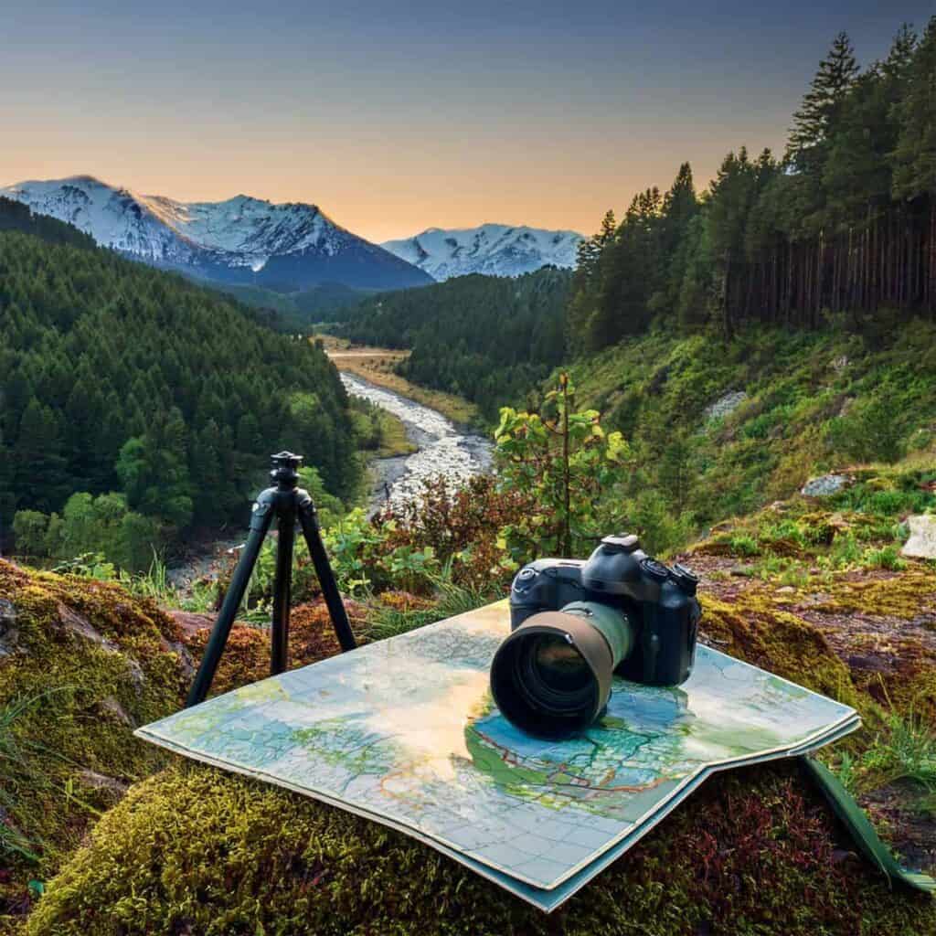 Camera and map on moss with a forest and mountain landscape in the background at sunrise or sunset.