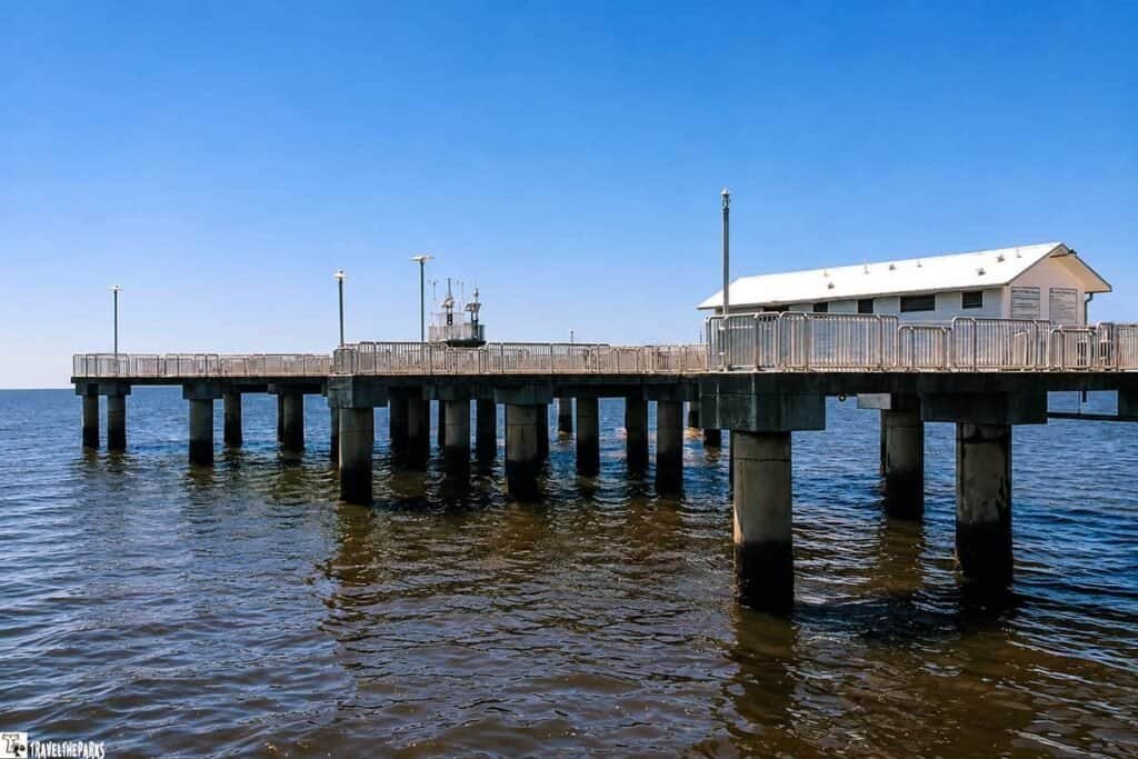 A concrete pier extends over calm waters, featuring a white building at the end and light poles along the sides under a clear blue sky.