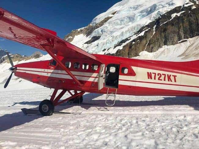 Flightseeing Denali K2 Air: A red and white airplane with skis parked on a snowy landscape in front of snowy mountains.

